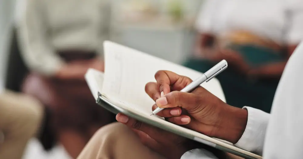 Close-up of a person writing in a notebook with a pen at Monumé Psychology assessment clinic, with others blurred in the background—an environment ideal for gifted testing and IQ assessment or learning and ADHD testing.
