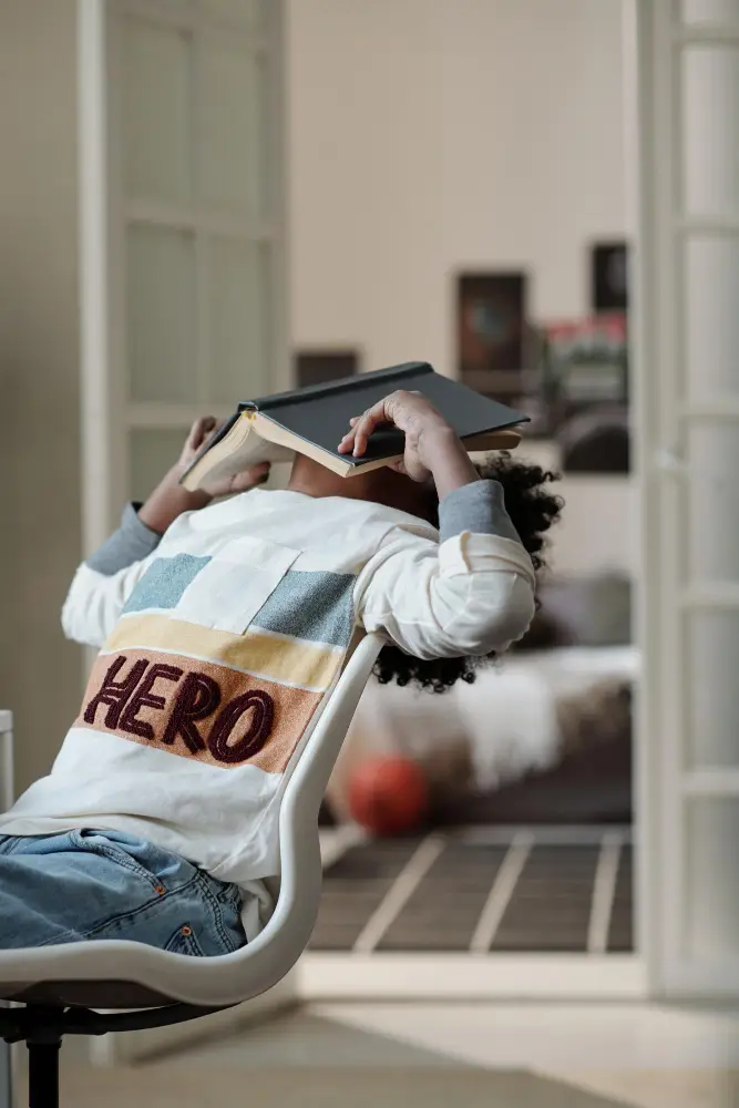 A child leans back in a chair with a book covering their face, wearing a sweatshirt that says "HERO," in a room with a bed and basketball—capturing the curiosity often explored at Monumé Psychology assessment clinic.