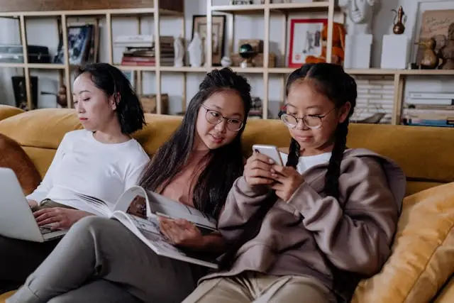 Three women sit on a yellow sofa; one uses a laptop, another reads a magazine about psychological assessment Mississauga, and the third checks her smartphone. Books and decor items are visible on shelves in the background.