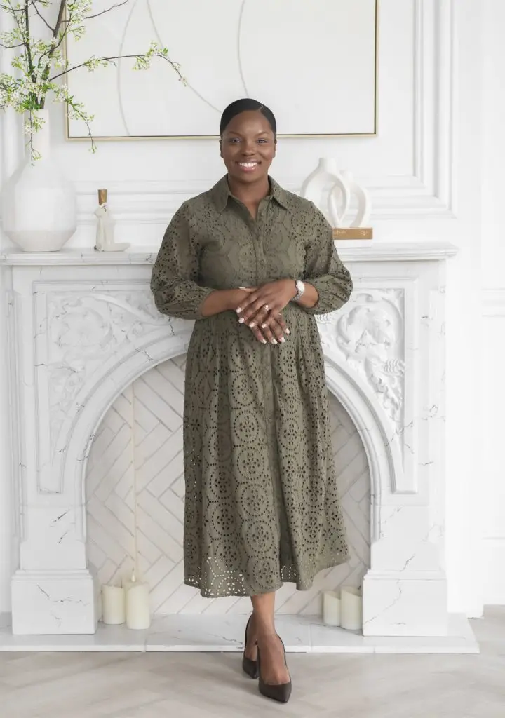 Sabrina McHugh-Thomas, Registered Psychologist & Founder of Monumé™ Psychology in a long, olive green eyelet dress stands in front of a white ornate fireplace at Monumé Psychology Assessment Clinic, where candles and decorative items line the mantel.