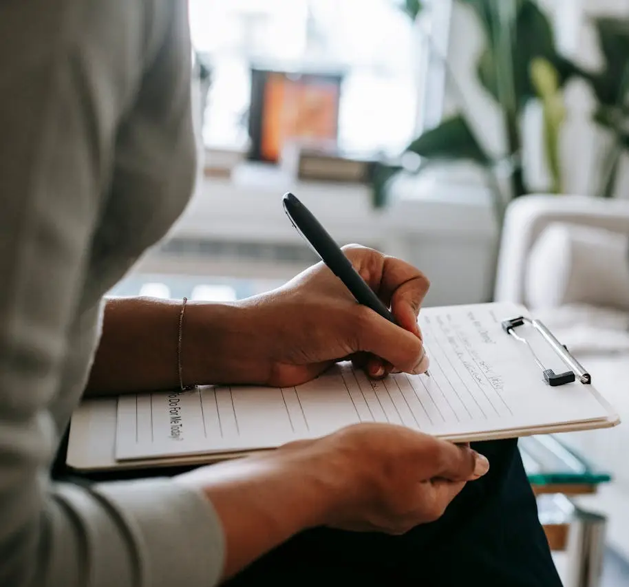A person writes on a clipboard with a pen, holding the paper steady with one hand. The blurred background features indoor plants and home decor, creating a calming setting for psychological assessment Mississauga or adult ADHD assessment and diagnosis.