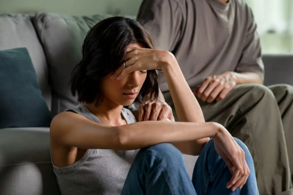 A woman sits on the floor with her hand on her forehead, appearing distressed, while a person on a couch rests a comforting hand on her shoulder—reflecting the support found at Monumé Psychology assessment clinic for learning and ADHD testing.