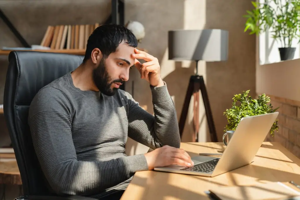 A man sits at a desk with a laptop, appearing focused and concerned, touching his forehead with one hand. A plant and lamp sit on the desk in this bright room at Monumé Psychology assessment clinic in Mississauga.