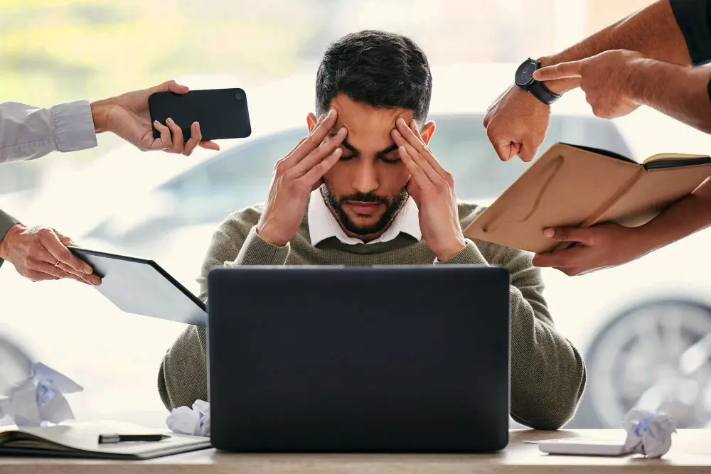 A man sits at a desk with his hands on his temples, looking stressed, while multiple hands offer him a phone, tablet, notebook, and point at his laptop—signs he may benefit from psychological assessment Mississauga or learning and ADHD testing.